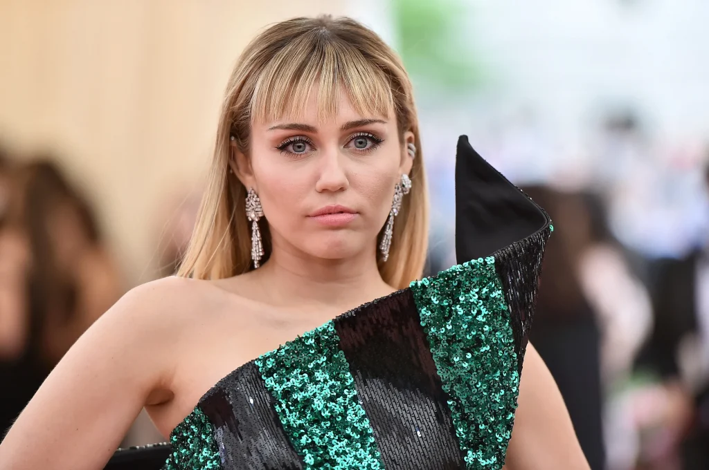 Miley Cyrus in off shoulder black and green sequence dress posing for camera and showing her Bangs and Blonde Hair - hairstyles of bollywood actresses