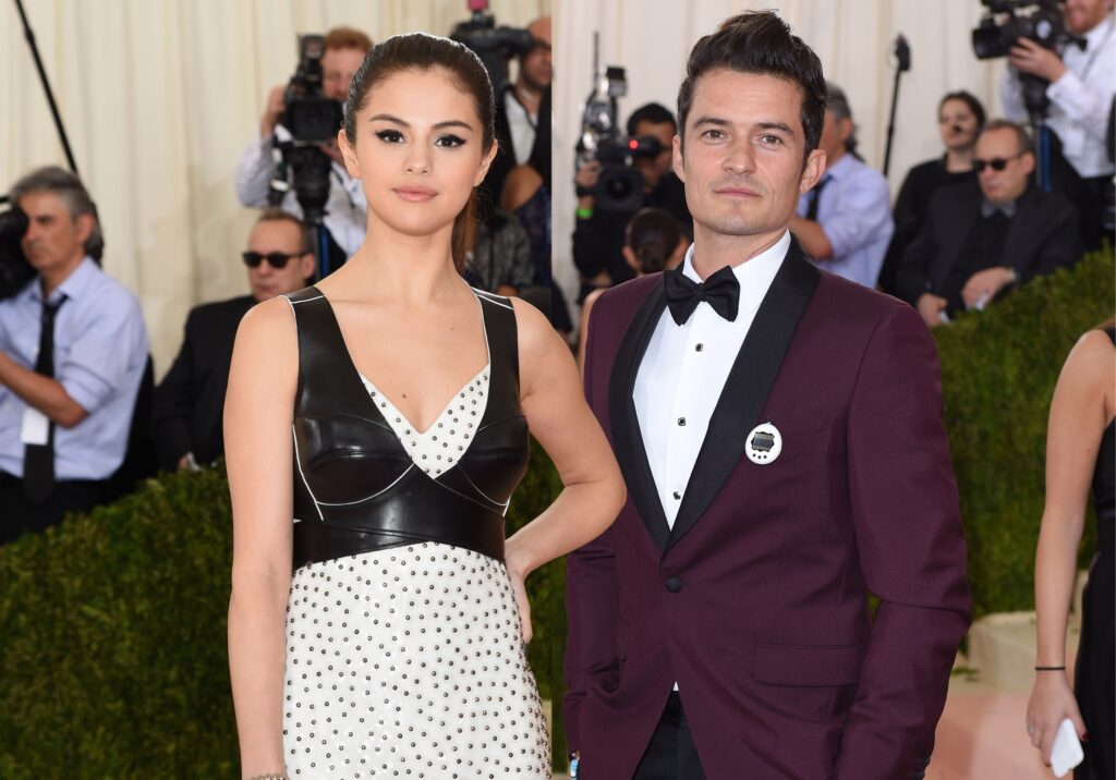 Selena Gomez in white polka dotted dress with black waste coat and Orlando Bloom in purple suit with white shirt posing for camera - selena gomez age