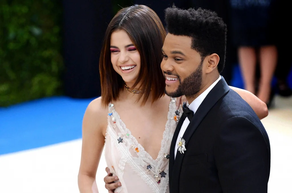 Selena Gomez in white deep neck dress and Weeknd in blue suit with white shirt posing for camera - selena gomez husband
