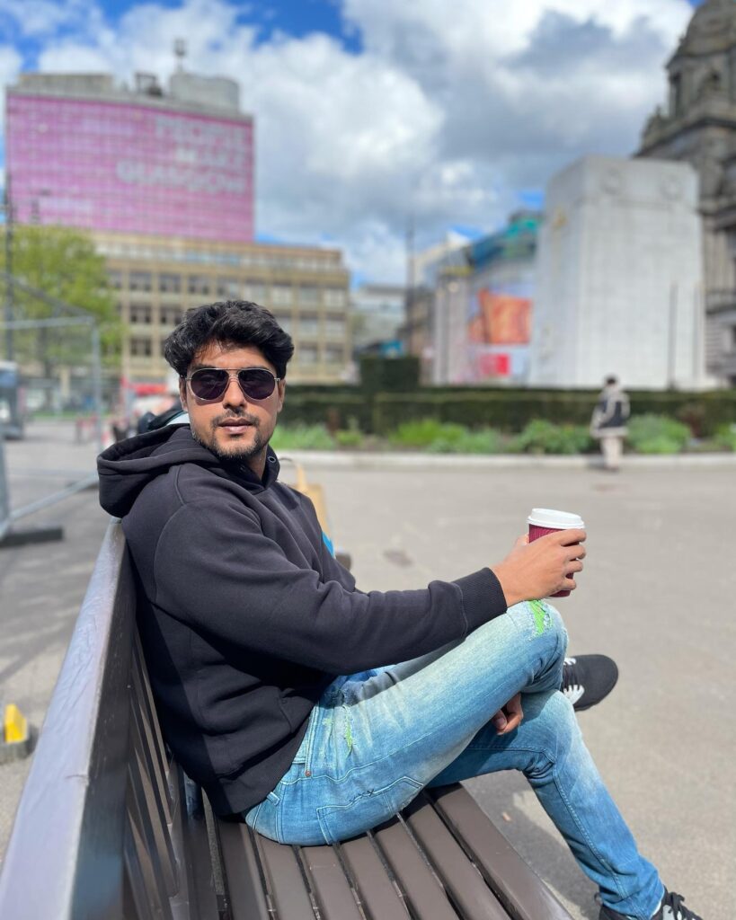 Ankit Gupta in black hoodie with blue jeans  and goggles sitting on a bench holding a cup of coffee - most handsome tv actors indian