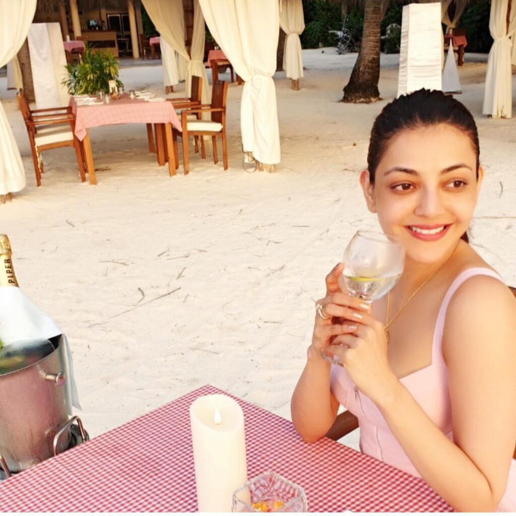 Smiling Kajal Agarwal sitting on a chair with a glass of drink in pink strappy dress - south actress without makeup