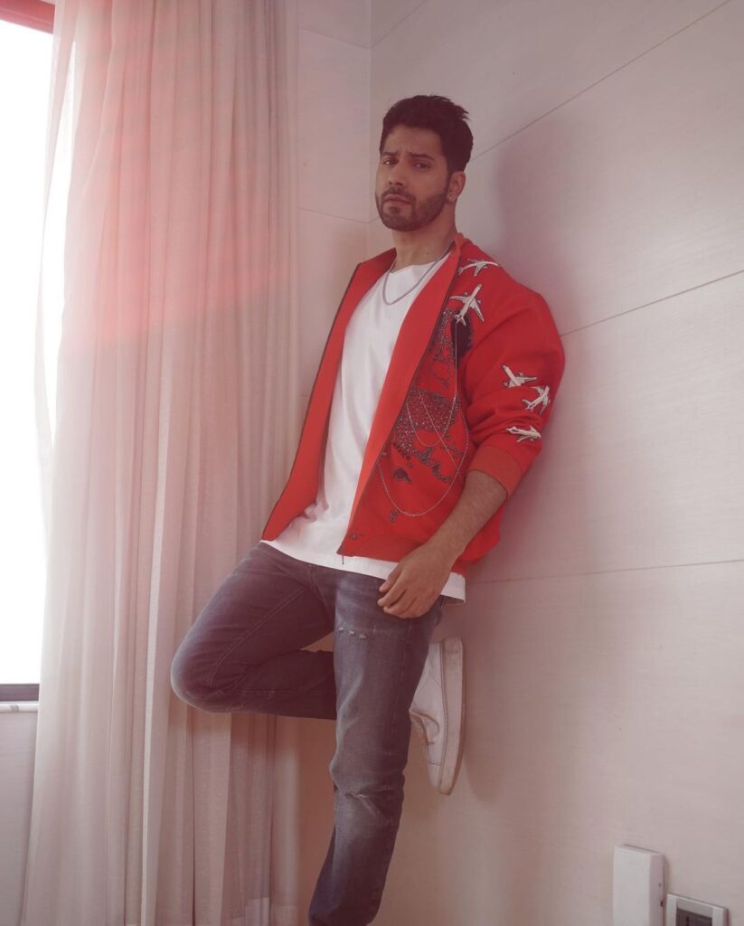 Varun Dhawan in red jacket with white t-shirt and blue jeans posing for camera - Varun Dhawan hair look